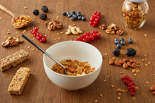 Jar and bowl with granola next to berries, nuts, cereal bars on wooden background