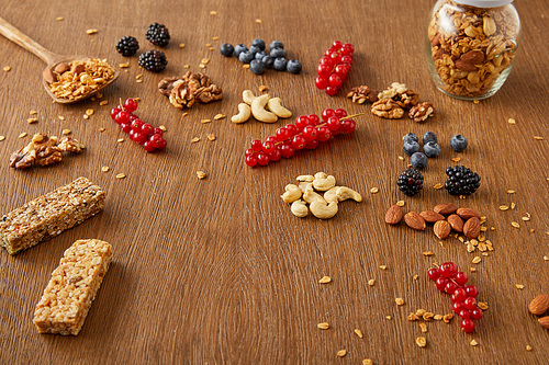 Jar of granola next to berries, nuts, cereal bars on wooden background