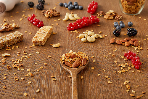 Wooden spatula with granola next to berries, nuts, oat flakes and cereal bars on wooden background