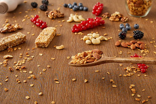 Wooden spatula above table with granola next to berries, nuts and cereal bars on wooden background