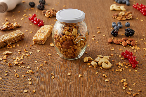 Selective focus of jar of granola with nuts, oat flakes, berries and cereal bars on wooden background