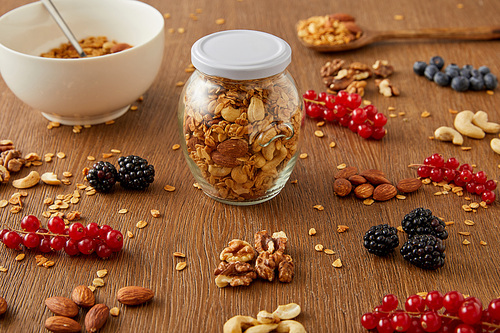 Selective focus of jar of granola and bowl next to nuts, oat flakes and berries on wooden background