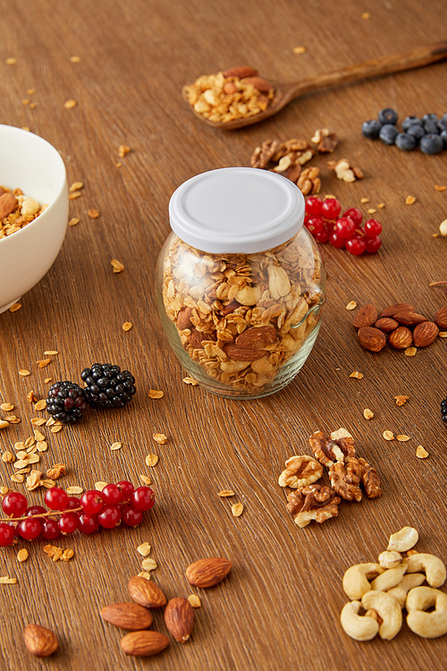 Jar of granola, bowl and spatula next to almonds, walnuts, cashews, berries on wooden background