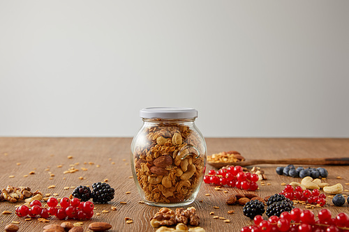 Selective focus of jar of granola with nuts, oat flakes, berries and cereal bars on wooden background isolated on grey