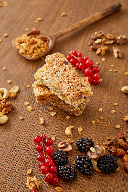 Selective focus of cereal bars, spatula with granola, redcurrants, blackberries and nuts on wooden background