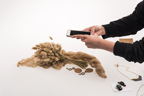 cropped view of photographer taking pictures of composition with flora and jewelry on smartphone on white