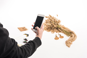 cropped view of commercial photographer taking pictures of composition with flora and jewelry on smartphone on white