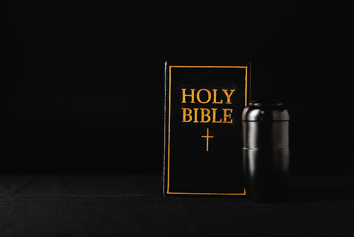 holy bible and urn with ashes on black background, funeral concept