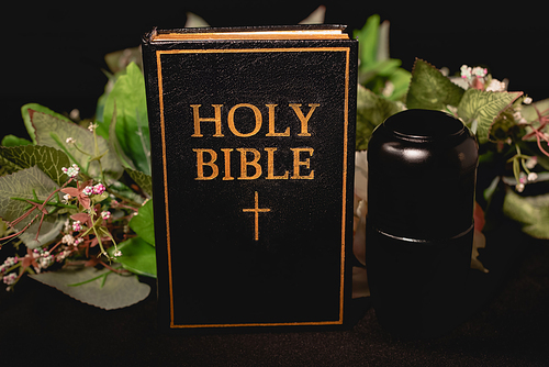 holy bible and urn with ashes on black background, funeral concept