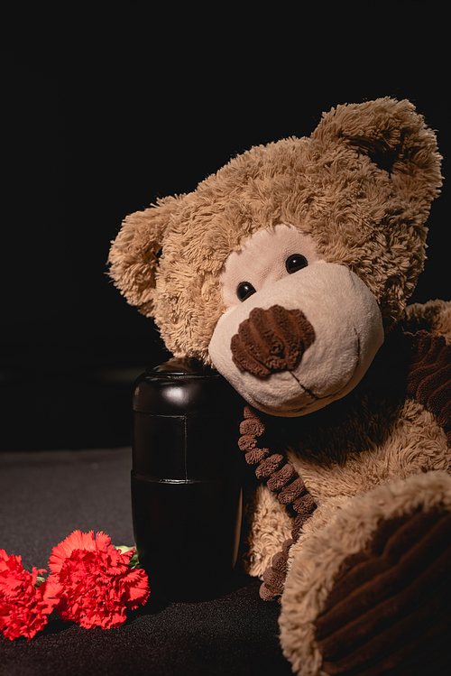 carnation, teddy bear and urn with ashes on black background, funeral concept