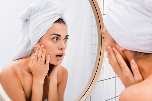 shocked young woman with white towel on head touching face while looking in mirror, blurred foreground