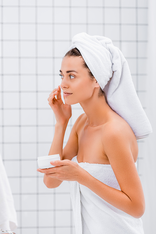 young woman with towel on head applying face cream in bathroom