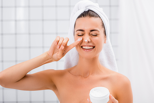 cheerful woman with closed eyes applying face cream on nose in bathroom