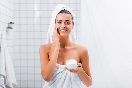 happy young woman, wrapped in towels, applying face cream and smiling at camera in bathroom