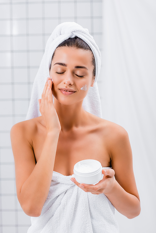 joyful young woman with closed eyes applying cosmetic cream on face in bathroom