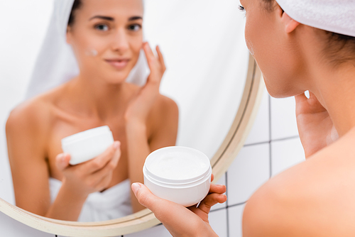 young woman applying face cream near blurred mirror reflection in bathroom