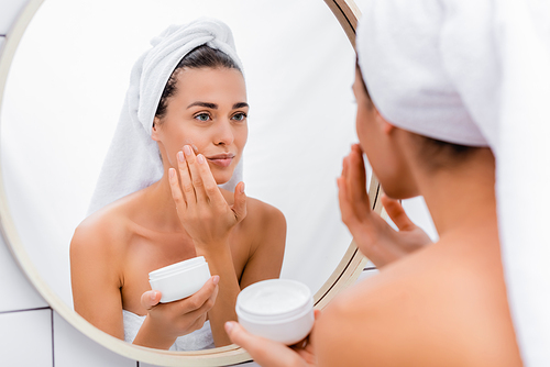 woman with white towel on head looking in mirror while applying face cream in bathroom