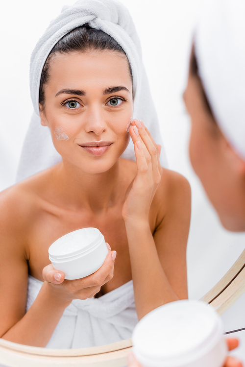 young woman with white towel on head applying face cream near mirror in bathroom, blurred foreground