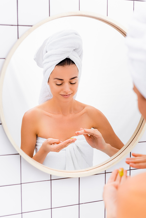 young woman with towel on head and facial scrub on hands near mirror in bathroom, blurred foreground