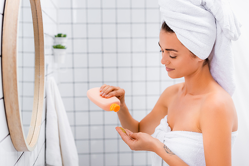 smiling woman with white towel on head holding bottle with facial scrub in bathroom