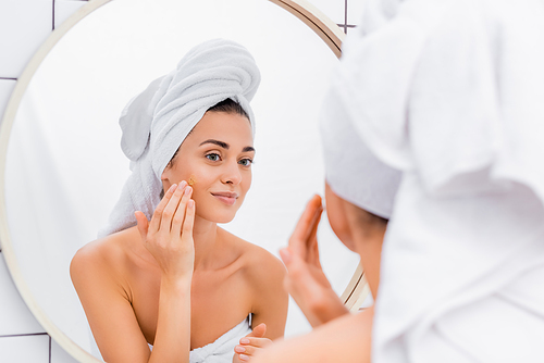 young woman with white towel on head applying facial scrub in bathroom, blurred foreground