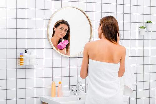 young woman, wrapped in white terry towel, drying hair near mirror in bathroom