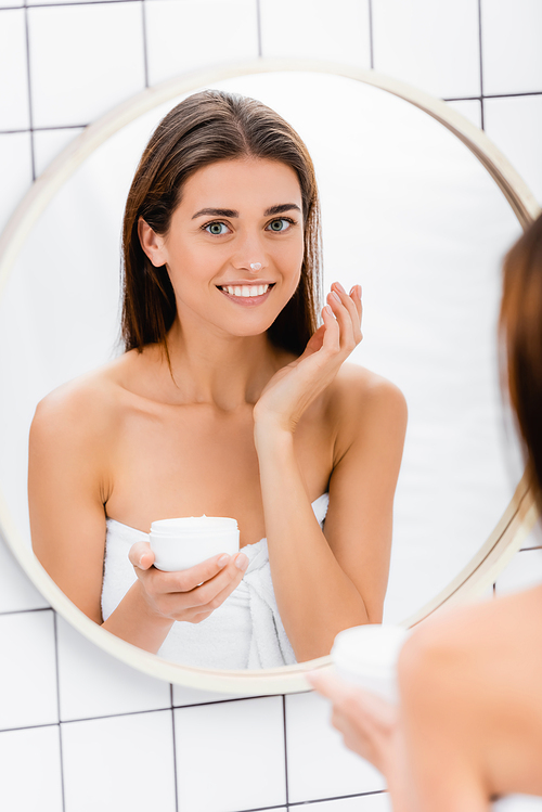 happy woman smiling while applying face cream near mirror in bathroom, blurred foreground