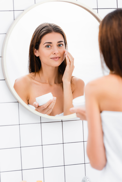 pleased woman applying face cream near mirror in bathroom, blurred foreground