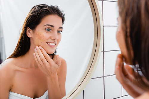 smiling woman looking in mirror while applying scrub on face in bathroom, blurred foreground