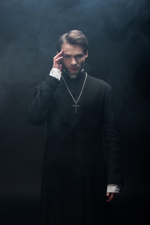young thoughtful catholic priest touching head while  on black background with smoke