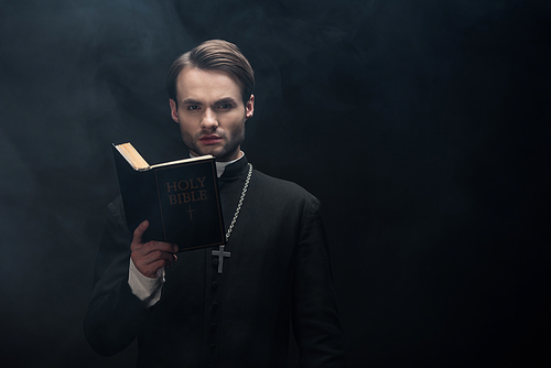 young serious catholic priest  while reading bible on black background with smoke