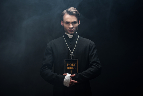 young confident catholic priest  while holding holy bible on black background with smoke