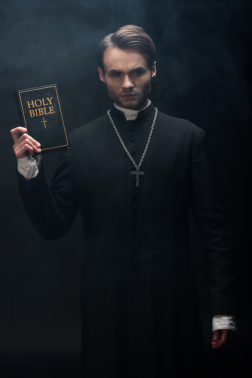 young strict catholic priest showing holy bible at camera on black background with smoke