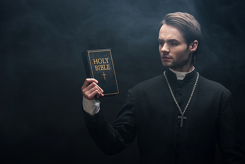 thoughtful catholic priest looking at holy bible on black background with smoke