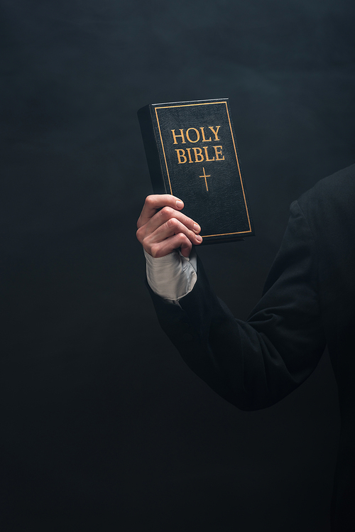partial view of catholic priest holding holy bible isolated on black