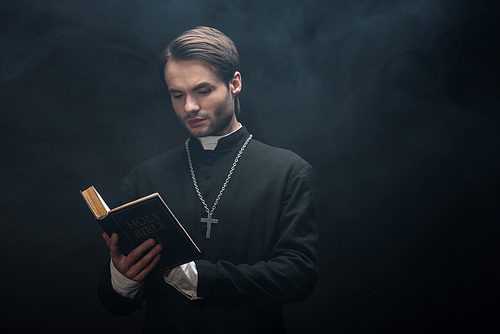 attentive catholic priest reading holy bible on black background with smoke