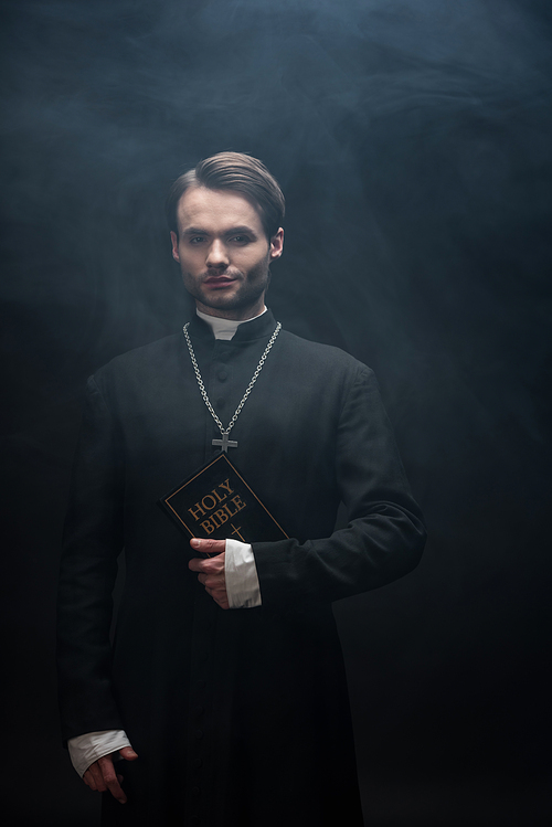 confident catholic priest holding holy bible and  on black background with smoke