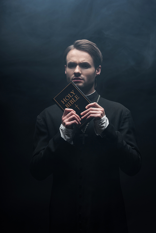 tense catholic priest  while holding holy bible on black background with smoke