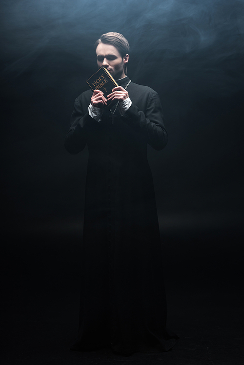 full length view of young catholic priest kissing holy bible on black background with smoke