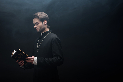 concentrated catholic priest reading holy bible on black background with smoke