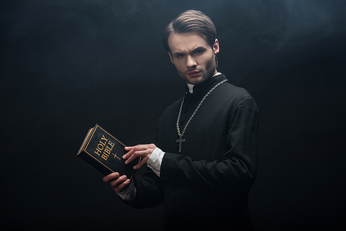 strict catholic priest holding holy bible and  on black background with smoke