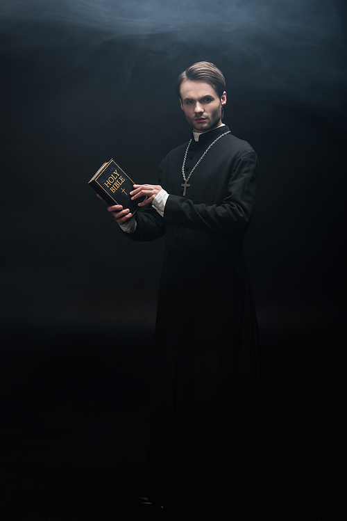 full length view of strict catholic priest holding holy bible and  on black background with smoke