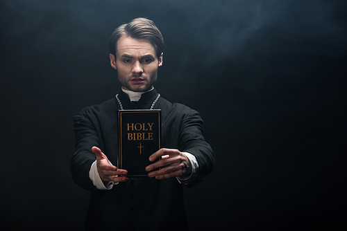 confident catholic priest holding holy bible in outstretched hand on black background with smoke