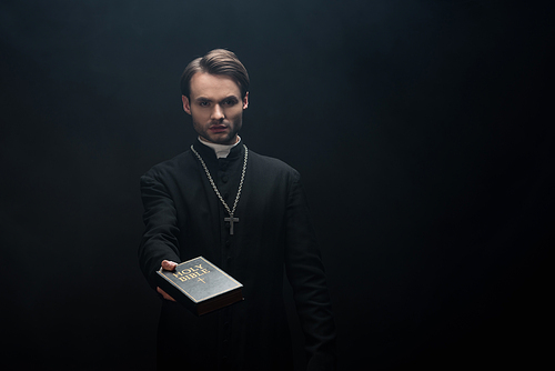 young serious catholic priest holding holy bible in outstretched hand isolated on black