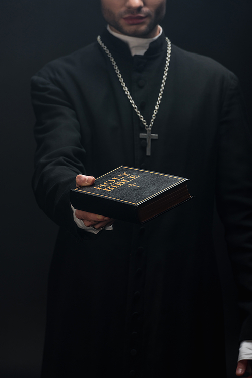 cropped view of catholic priest holding holy bible in outstretched hand isolated on black
