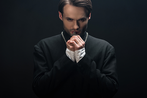 young thoughtful catholic priest holding silver cross on his necklace isolated on black