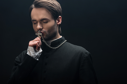 young thoughtful catholic priest kissing silver cross with closed eyes isolated on black
