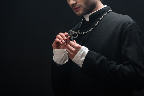cropped view of catholic priest looking at silver cross on his necklace isolated on black