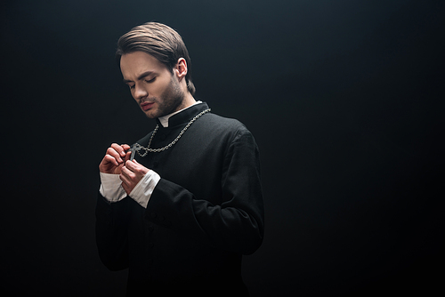 young serious catholic priest looking at silver cross on his necklace isolated on black