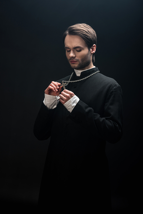 young thoughtful catholic priest looking at silver cross on his necklace isolated on black
